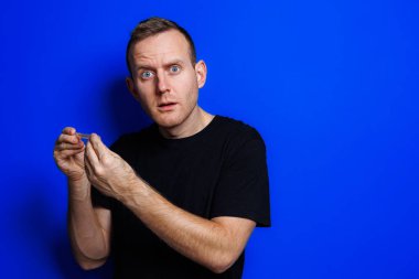 A young man in a black T-shirt on a blue background stands with a thermometer in his hands. An increase in body temperature, feeling sick. Weakened immunity. Selective focus