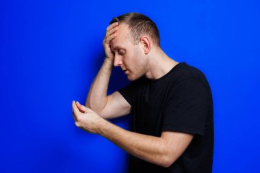 A young man in a black T-shirt on a blue background stands with a thermometer in his hands. An increase in body temperature, feeling sick. Weakened immunity. Selective focus
