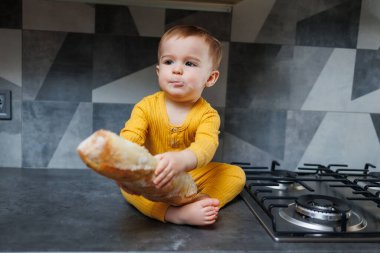 A cute 1-year-old boy in yellow clothes is sitting in the kitchen on the table with a fresh wheat baguette. Portrait of a cute one-year-old boy eating delicious bread