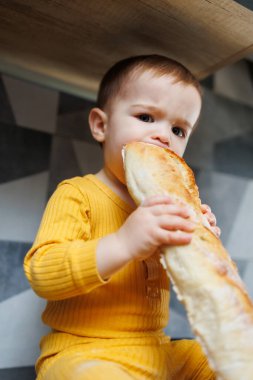 Bir yaşındaki küçük bir çocuk mutfakta oturuyor ve mutfakta uzun bir ekmek ya da ekmek yiyor..