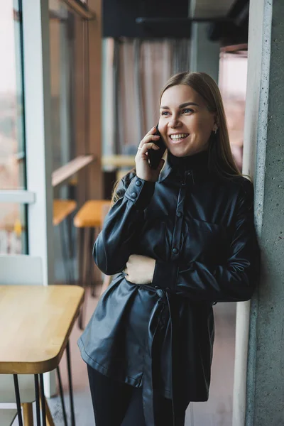 A smiling young woman is standing with a mobile phone in her hands and looking to the side while standing near a window reflecting sunlight. Modern workspace. Woman freelancer