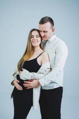 Happy young man standing with his pregnant wife and hugging her belly, isolated on white background. Stylish young people. A man in a shirt and a woman in a black skirt and jacket