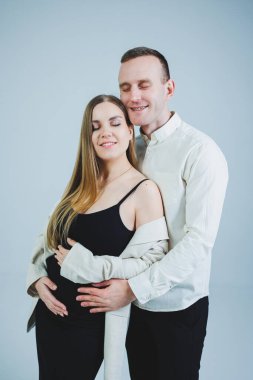 Happy young man standing with his pregnant wife and hugging her belly, isolated on white background. Stylish young people. A man in a shirt and a woman in a black skirt and jacket
