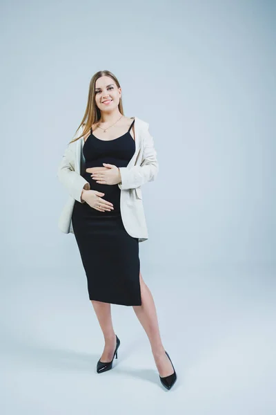 A young woman in a black top, skirt and beige jacket stands on a white background and hugs her pregnant belly. Attractive pregnant woman on a white background. Casual clothes for stylish and fit women