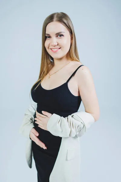 A young woman in a black top, skirt and beige jacket stands on a white background and hugs her pregnant belly. Attractive pregnant woman on a white background. Casual clothes for stylish and fit women