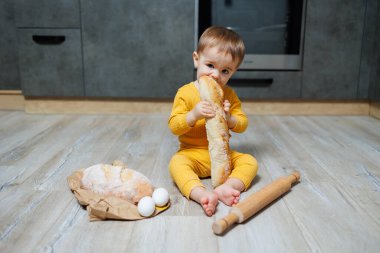 A cute little boy one year old sits and eats freshly baked rye bread. The child holds a fresh baguette in his hands.