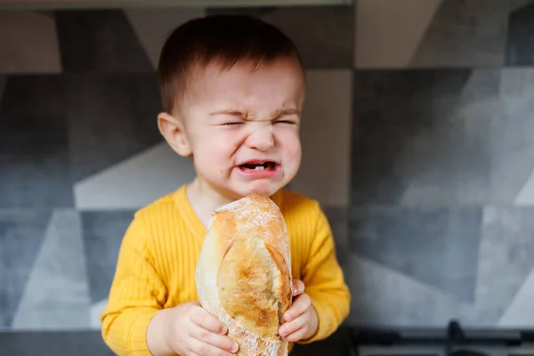 A one-year-old little boy in yellow clothes sits and eats freshly baked rye bread. The child holds a fresh baguette in his hands.