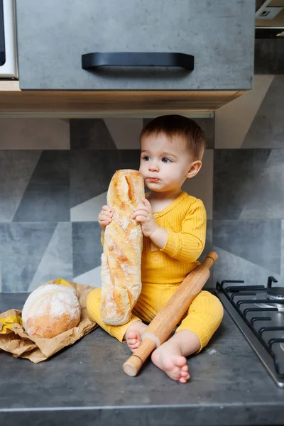 A cute little boy one year old sits and eats freshly baked rye bread. The child holds a fresh baguette in his hands.