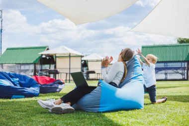Mother with laptop, her son playing outdoors on green lawn. Rest and work with a child in the park