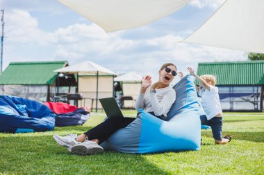 Mother with laptop, her son playing outdoors on green lawn. Rest and work with a child in the park
