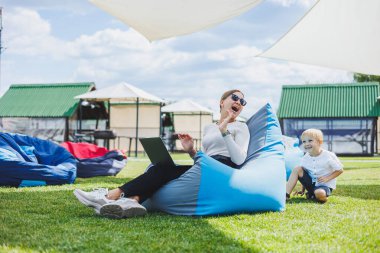 Mother with laptop, her son playing outdoors on green lawn. Rest and work with a child in the park