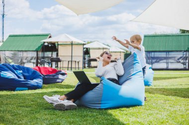 Mother with laptop, her son playing outdoors on green lawn. Rest and work with a child in the park
