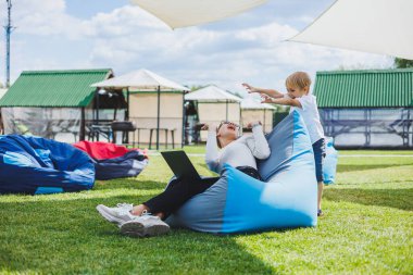 Mother with laptop, her son playing outdoors on green lawn. Rest and work with a child in the park