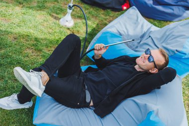A young man sits in a chair outdoors and smokes a hookah,