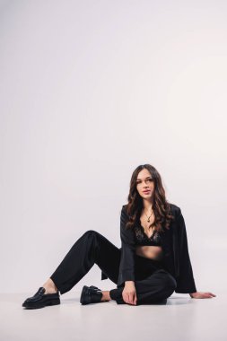 A stylish pretty young woman in a black classic suit is sitting on a white background. A beautiful woman in a jacket and make-up on her face