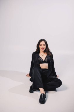 A stylish pretty young woman in a black classic suit is sitting on a white background. A beautiful woman in a jacket and make-up on her face