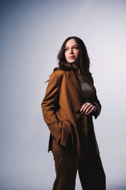 Stylish pretty young woman in brown classic suit sitting on gray background. Beautiful woman in a jacket and with makeup on her face