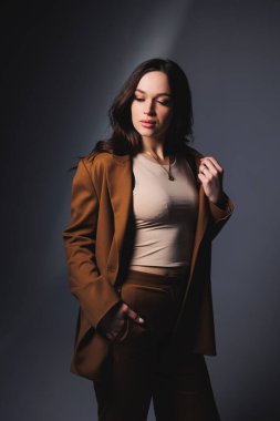 Stylish pretty young woman in brown classic suit sitting on gray background. Beautiful woman in a jacket and with makeup on her face