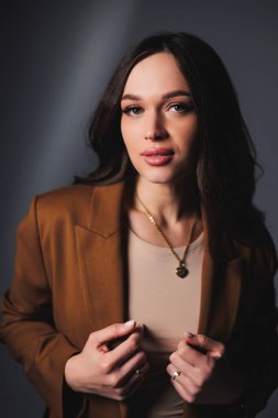 Stylish pretty young woman in brown classic suit sitting on gray background. Beautiful woman in a jacket and with makeup on her face