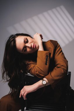 A pretty young woman with a smile on her face and wearing a brown classic suit is sitting on a chair on a gray background. Beautiful woman in a jacket and with makeup on her face