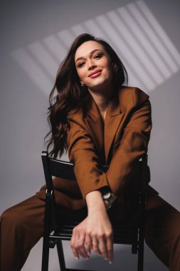 A pretty young woman with a smile on her face and wearing a brown classic suit is sitting on a chair on a gray background. Beautiful woman in a jacket and with makeup on her face