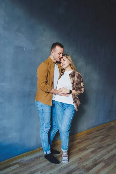 A young married couple in love in shirts and jeans on the background of a gray wall. The concept of happy family relationships. A man and a woman are hugging