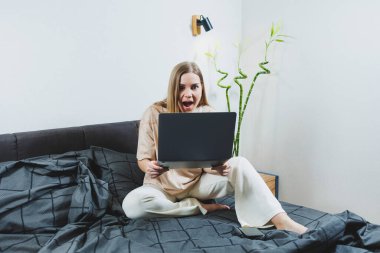 Smiling young woman in casual clothes smiling and working remotely, sitting on bed with laptop during remote work. Freelance work. Work from home