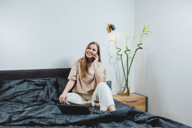Smiling young woman in casual clothes smiling and working remotely, sitting on bed with laptop during remote work. Freelance work. Work from home