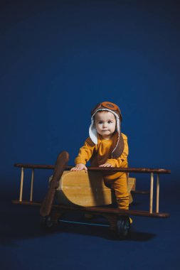 A little boy of 1 year in a yellow knitted suit and an aviator hat stands near a wooden helicopter. Children's wooden ecological toys made of antural wood