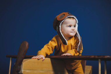 A little boy of 1 year in a yellow knitted suit and an aviator hat stands near a wooden helicopter. Children's wooden ecological toys made of antural wood