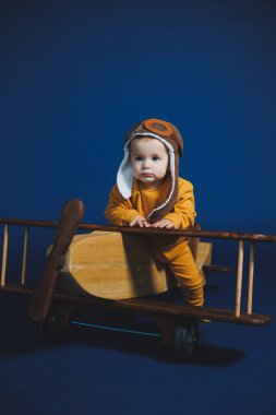 A little boy of 1 year in a yellow knitted suit and an aviator hat stands near a wooden helicopter. Children's wooden ecological toys made of antural wood