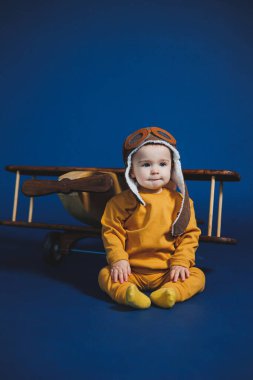 A little boy of 1 year in a yellow knitted suit and an aviator hat stands near a wooden helicopter. Children's wooden ecological toys made of antural wood