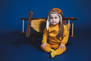 A little boy of 1 year in a yellow knitted suit and an aviator hat stands near a wooden helicopter. Children's wooden ecological toys made of antural wood