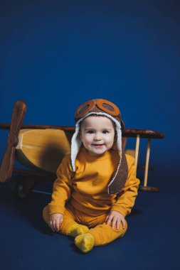 A little boy of 1 year in a yellow knitted suit and an aviator hat stands near a wooden helicopter. Children's wooden ecological toys made of antural wood