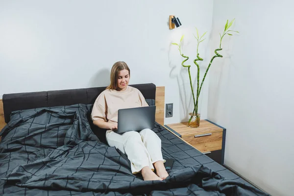 Satisfied young woman finishing work on laptop. European girl sitting with computer in bed, studying at home, distance learning and remote freelance work concept