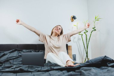 Satisfied young woman finishing work on laptop. European girl sitting with computer in bed, studying at home, distance learning and remote freelance work concept