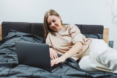 Satisfied young woman finishing work on laptop. European girl sitting with computer in bed, studying at home, distance learning and remote freelance work concept