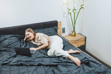 Satisfied young woman finishing work on laptop. European girl sitting with computer in bed, studying at home, distance learning and remote freelance work concept