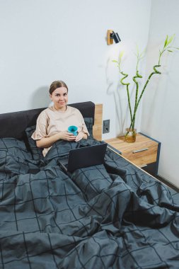 Young beautiful woman sitting on bed with coffee, working with computer at home, remote work. Woman happy and smiling happy in bed at home.