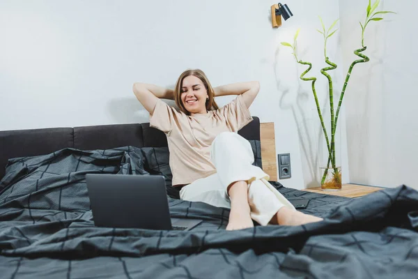 Satisfied young woman finishing work on laptop. European girl sitting with computer in bed, studying at home, distance learning and remote freelance work concept