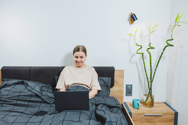 Молодая красивая женщина сидит на кровати с кофе, работает с компьютером дома, удаленная работа. Женщина счастлива и улыбается счастливо в постели дома.