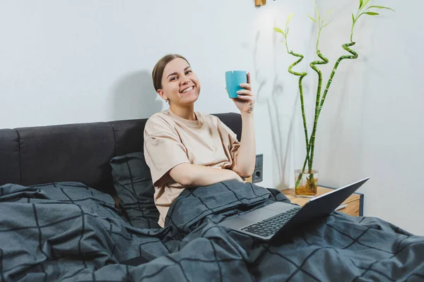 stock image Young European beautiful woman sitting on bed with coffee, working with computer at home, remote work. Woman happy and smiling in bed at home.