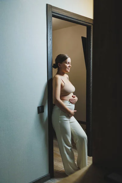 Cute young pregnant woman in a top and casual maternity clothes. Cute pregnant woman hugging her belly at home