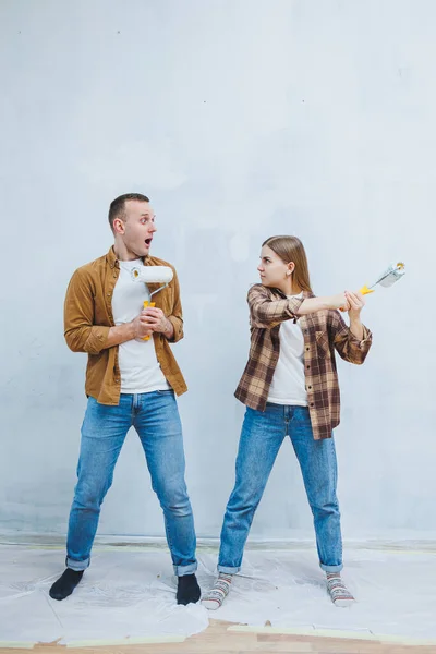 stock image Happy young married couple in love in shirts doing renovations, renovating walls painting with white paint roller, preparing to move into new house, selective focus.