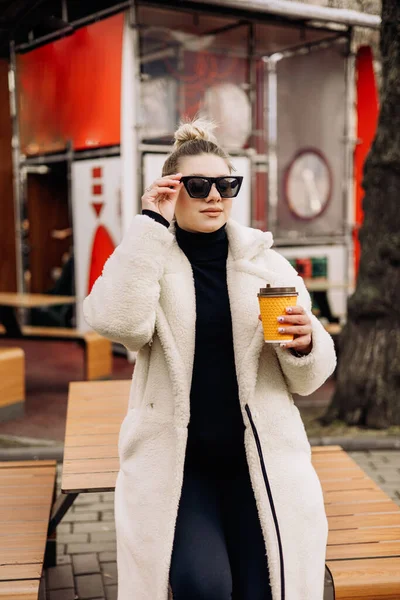 白い毛皮のコートを着た若いきれいな女性とコーヒーを片手にサングラスが街を歩き回っています スタイリッシュな若い女の子 — ストック写真