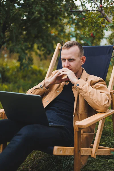 stock image Cute man in a shirt sitting in a chair outdoors in the garden and working on a laptop, remote work, female freelancer