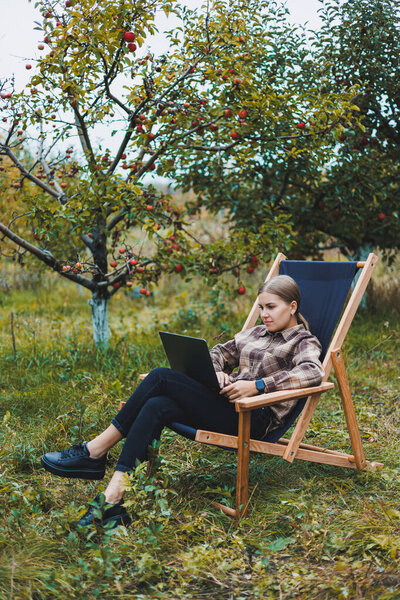 Beautiful woman in checkered shirt with laptop working outdoors in garden, home office concept. Remote work.