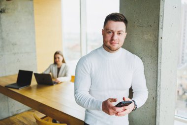 Gündelik giysiler içinde başarılı bir erkek işadamı modern ışık ofisinde telefonuyla telefonuna bakarak gülümsüyor..