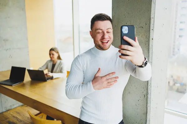 stock image Positive male businessman in sweater smiling looking at phone standing in modern light office with phone working in modern company.