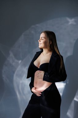 Siyah klasik takım elbiseli hamile bir kadının portresi. Gri arka planda hamile bir kadın karnına sarılıyor..